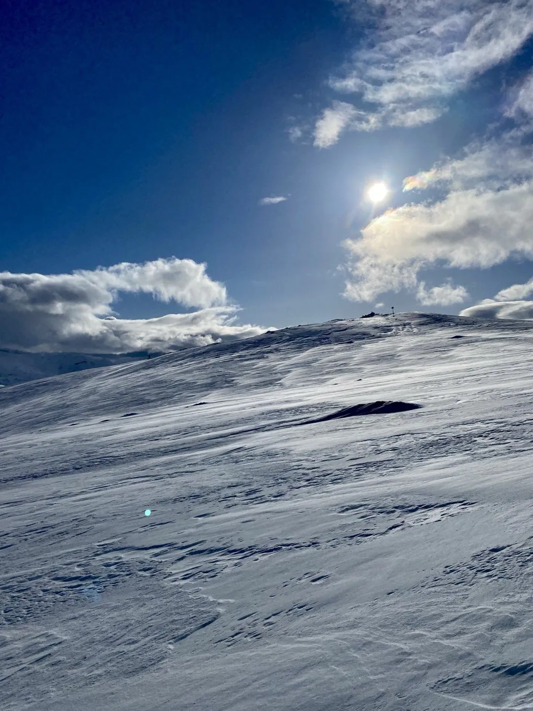 Vindblåst snø på et fjellplatå under blå himmel, med sol og skyer